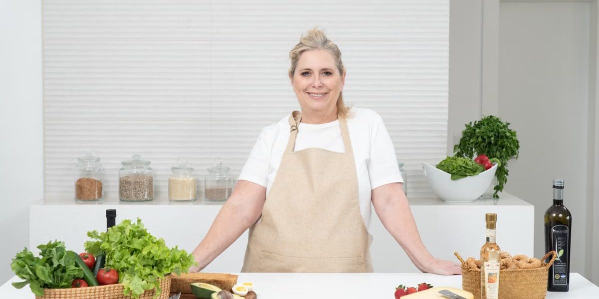 A mature woman in an apron stands at a kitchen counter with fresh produce and cheese, ready for cooking.