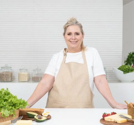 A mature woman in an apron stands at a kitchen counter with fresh produce and cheese, ready for cooking.