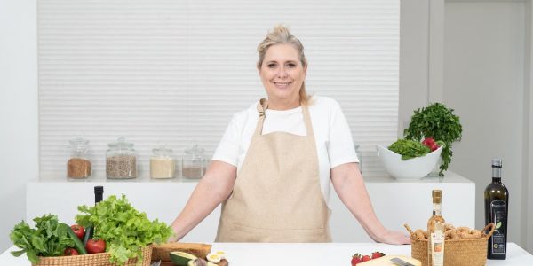 A mature woman in an apron stands at a kitchen counter with fresh produce and cheese, ready for cooking.